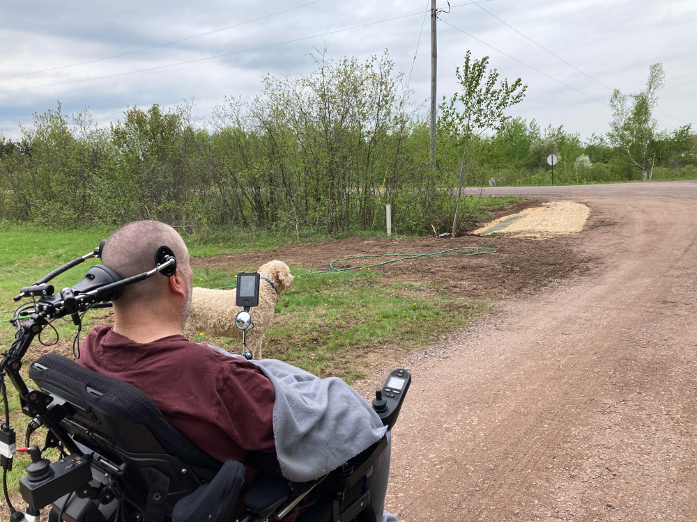 ALS progression | ALS News Today | A photo is taken from behind Todd, who sits in his power wheelchair, inspecting work on the culvert in the couple's driveway, along with a recently planted tree.
