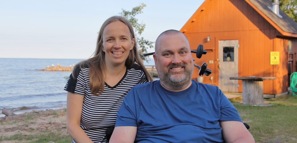 life with ALS | ALS News Today | A scenic photo of Todd and Kristin in front of Lake Superior, in northern Michigan, in 2020. An orange cabin is visible in the background