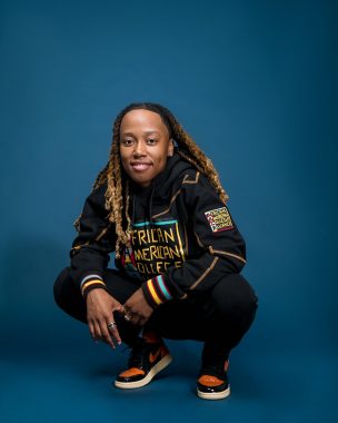 A portrait photo of Aleia Kristene, in which she is squatting before an all-blue background and floor. She is smiling and wearing black clothing with blue and yellow writing and white, black, and orange sneakers. 