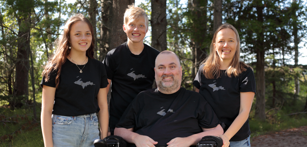 living with als | ALS News Today | A photo of the Neva family in the woods of northern Michigan. Todd is seated in his chair in the middle and flanked by his two children and Kristin. They're all wearing black T-shirts, and rays of bright sunshine filter through some of the majestic pines
