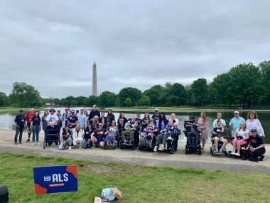 A group photo with about three dozen people in front of the Washington Monument and the Lincoln Memorial Reflecting Pool. In the foreground is a sign that says "I Am ALS."