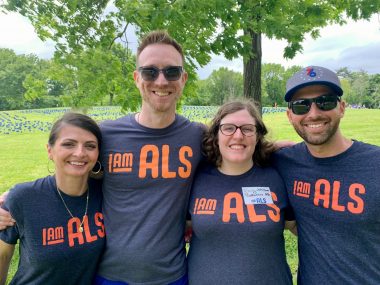 Two men and two women, all dressed in navy blue T-shirts with the words "I Am ALS" printed in orange, stand in a park and embrace for a group photo.