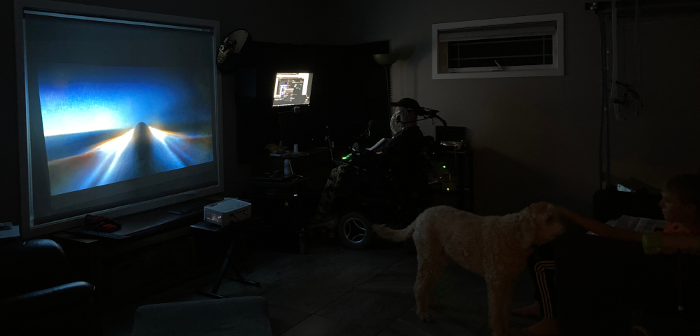A wide photo shows a dark room where the movie "Top Gun: Maverick" is being projected onto a large window shade. A man is sitting to one side in his wheelchair, and a dog and a kid are visible to the right. 