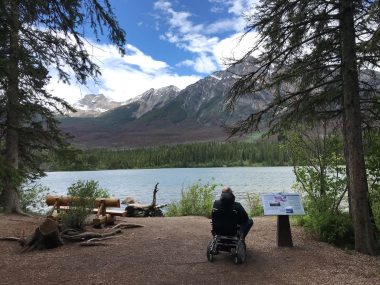 A man sits in a wheelchair looking out over a body of water at mountain peaks and a blue sky with clouds. On the bank with him are trees, a marker, and a bench.