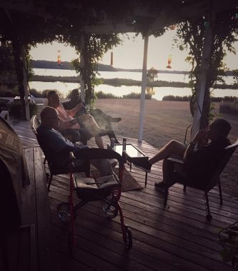 A shadowy filtered photo with vignette shows three men sitting on the back deck of a house overlooking a body of water; the men are visiting and smoking cigars