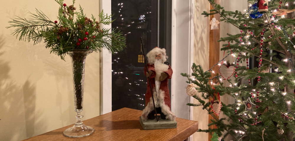 The top of a bookshelf features a Santa figure made from wood, burlap, leather, and wool. To the left is a tall glass Mikasa vase filled with pine sprigs and red berries. On the right is a balsam fir Christmas tree decorated with lights, ornaments, and beaded garland.