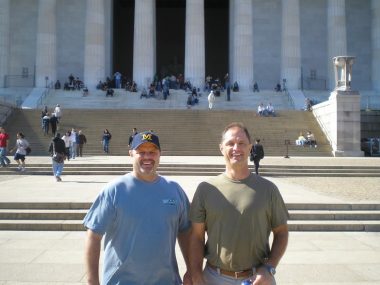 Two middle-aged brothers stand in front of the Lincoln Memorial in Washington, D.C. It's a sunny day and the two men are dressed in T-shirts. One is wearing a sports cap. 