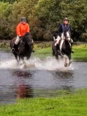 Two women wear riding gear and helmets as they ride horses through a shallow pond in Ireland. The horse on the left is dark brown, while the horse on the right is brown and white. The women appear to be laughing.