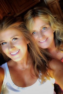 This photo features head-and-shoulders images of two blonde-haired women. The one on the left is in a white tank top and wears a subtle necklace; the other appears to be wearing an orange-red top.