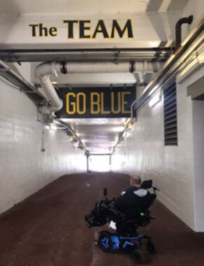 A man sits in his blue power wheelchair looking down a long hallway inside Michigan Stadium. "The TEAM" and "GO BLUE" are painted in blue and gold above him.