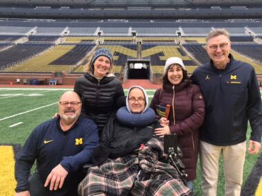 Five family members pose together on the 50-yard line at Michigan Stadium. In the center is Jeff, who's seated in his power wheelchair. Standing behind him to his right is his wife, Juliet. Another man kneels by Jeff's right side, and a man and a woman stand to his left.