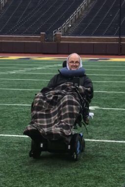 A man guides his blue power wheelchair down the field at the University of Michigan stadium. He has a flannel blanket draped across his lap and a blue pillow supporting his neck.
