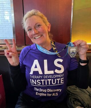 This photo shows a woman with tied-back blonde hair displaying the peace sign with one hand while holding up a medal with the other. She's indoors and wears a purple T-shirt with the words "ALS Therapy Development Institute" and "The Drug Discovery Engine for ALS." 
