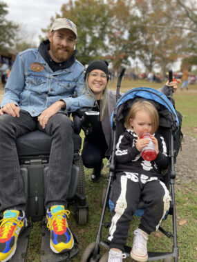 A family photo in a park shows a young husband in his mid-30s, wearing a jean jacket, tan baseball cap, and colorful sneakers, and in a power wheelchair, next to his wife, who's head is visible from behind, and their young son, who is dressed in a skeleton outfit and is sitting in a stroller. 