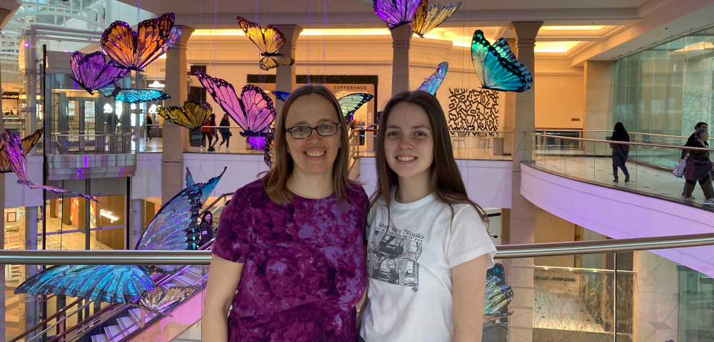A woman and a teenager stand in the upstairs of a mall. Behind them are columns and decorative butterflies. The woman has brownish-blond hair and wears glasses and a purple print shirt. The teenager has long brown hair and wears a white T-shirt with a design in black.