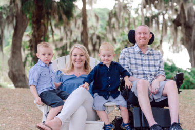 A family portrait taken outdoors, with trees and a body of water in the background. Pictured are two young boys and a husband and wife. The husband is in a power wheelchair. It is warm out, as all of the boys are wearing shorts.