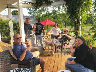 A photo of a beautiful back deck to a house. The deck sits among trees, plants, and other greenery that seems to stretch to the horizon. On the deck are six middle-aged people in various stages of relaxation, drinking a beer, smoking a cigar, etc. The sun is shining and the photo projects a sense of happiness.