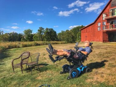 A man reclines in his power wheelchair on the grassy slopes of the South River. Two patio chairs are just to his right, placed side by side, and a large red house resembling a barn is in the background. It appears to be a warm, sunny day.
