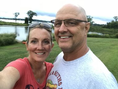 A husband and wife take a selfie while standing in their backyard at their home in Maryland. The woman is wearing a pink shirt and has sunglasses resting on her head, while the man is wearing a gray shirt and glasses. Several bodies of water are visible in the background.