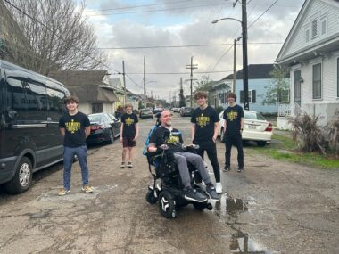 A father poses outside on a street with his four sons. He's sitting in a wheelchair, and they're standing and spaced out behind him.