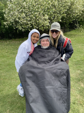 Three people pose outside on a short-trimmed lawn in front of some bushes. One is a young girl, another is a man in a wheelchair and covered by a blanket, and the third is a woman wearing a hat and dark sunglasses. The girl is wearing a hoodie covering most of her head. 