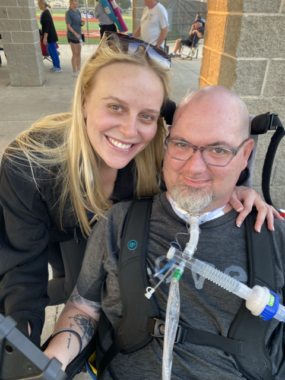 A smiling woman with long, blond hair and sunglasses perched on the top of her head leans forward with her left arm around a bald man with aa goatee, who is wearing glasses and in a power wheelchair. He is wearing all black, tattoos are partially visible on his right arm, and he has a tracheostomy. He also is smiling. Both people are looking at the camera. They appear to be outdoors somewhere, perhaps a football field or track. 