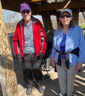 A man and a woman stand next to each other underneath some type of raised platform or other structure on an outdoor walking trail. Both sun and shadows are cast across the two. The man is wearing a purple baseball cap, sunglasses, and and a red jacket, the woman, a blue baseball cap, blue shirt, and sunglasses. 