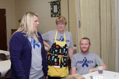 A woman wearing an apron stands between her son and his wife in front of a table at a fundraiser. They're all wearing gray shirts with a blue "Team Seth" logo.