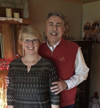 A couple poses for a photo inside their home. The woman has short blond hair and glasses and is wearing a black top and necklace. The man has graying hair and a mustache, and he's wearing a long-sleeved white shirt and a red vest. 