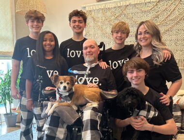 A family of seven poses for a photo with their two dogs. They're all wearing matching black-and-white plaid pajama pants and a black T-shirt with the word "love" printed in white. The father sits in the center in his wheelchair, with his wife, four sons, and daughter gathered around him.