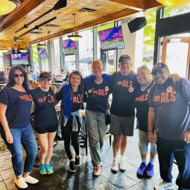 Seven people stand in a row with their arms around each other's shoulders. They're all wearing a T-shirt that says "I Am ALS" on the front in orange lettering, and they appear to be standing inside a restaurant.