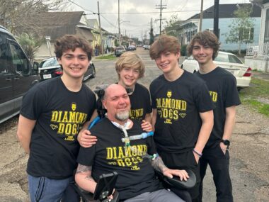 A father poses outside with his four sons for a photo. They're all wearing matching black shirts with a yellow "Diamond Dogs" logo on the front. The father is sitting in his wheelchair, with his trach visible, while his sons stand in a row behind him.