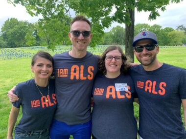 Two men and two women stand together with their arms around each other's shoulders. They're all wearing the same dark gray T-shirt with the orange "I Am ALS" logo on the front. In the background, thousands of blue flags representing people with ALS are planted on a sprawling lawn.