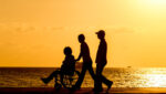 Three people, one in a wheelchair, walking on a beach at sunset