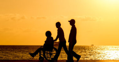 Three people, one in a wheelchair, walking on a beach at sunset