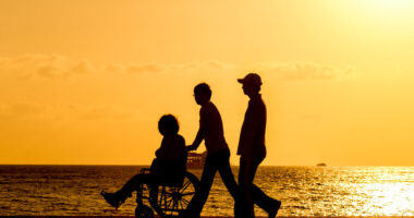 Three people, one in a wheelchair, walking on a beach at sunset