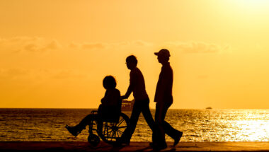 Three people, one in a wheelchair, walking on a beach at sunset