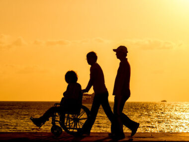 Three people, one in a wheelchair, walking on a beach at sunset