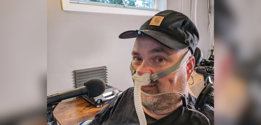 A closeup of a man wearing an AirFit CPAP nasal pillows mask and a black baseball cap. He is in a power wheelchair sitting in an office in front of a microphone.