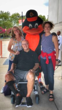 A vertical photo of two women flanking a man in a wheelchair. Between the two women is the Baltimore Oriole baseball team mascot. 
