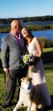 A newly married couple poses for a photo on their wedding day. The man is wearing a gray suit over a pink shirt, and the woman is wearing a sleeveless wedding dress and holding a bouquet of flowers. His head is turned to kiss his wife's forehead, and in his right hand, he holds the leash to the couple's husky mix, who sits at their feet.