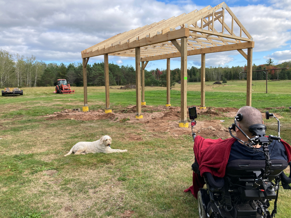 In the right foreground, a man in a red shirt sits in a wheelchair, his back to the camera. He is facing the assembled frame of a small structure, with four wooden poles on two sides and boards forming the frame of a roof. That all sits in a clearing of short grass, some of it worn. A light-colored dog lies down on the left, with what appears to be a red tractor and a stand of green trees in the background.