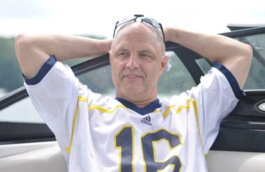 A middle-age man clasps his hands behind his head while leaning against a boat, with a blurry background showing sky, greenery, and water. He has sunglasses moved back atop his bald head and wears a white football jersey showing the number 16 in navy blue with a yellow outline.