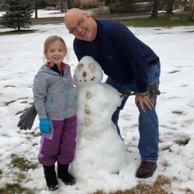 A man and his young niece, who could be 8 or 9, stand on either side of a small snowman they built. The man is leaning over, with his hands on his knees, to fit into the frame with the girl and the snowman. They're standing in a large yard that's mostly covered in snow, but a few grassy patches are visible under some trees in the background.