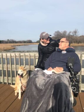 A man, woman, and dog pose on a wooden walkway in front of a lake. The couple looks each other in the face as the photo is taken. They're wearing winter clothing, and the man is sitting in a power wheelchair, covered in a blanket. Next to them is a dog sitting with his tongue out. It's a picturesque winter scene.