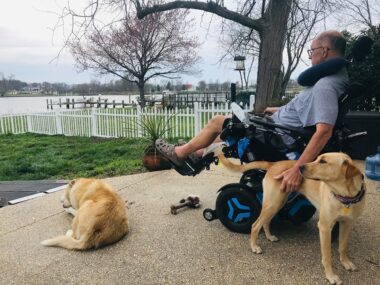 A man reclines in his power wheelchair on a patio behind his home. He's petting one dog with his left hand while another dog lies a few feet away, and looking out at a river.