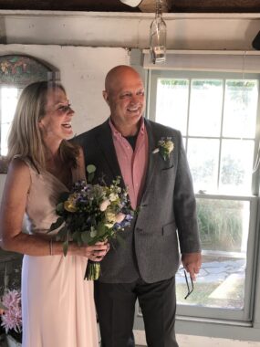 A couple stands side by side, both laughing, inside of a milk house on their rented property. They're dressed in wedding attire — the woman in a long white dress holding a bouquet, the man in a gray suit jacket and pink button-down shirt.