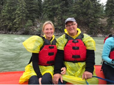 A man and a woman, both wearing yellow ponchos and red life jackets, sit on an orange raft while floating down a Canadian river. The water behind them has a green hue, and the riverbank is full of pine trees. It's a gray day, but the couple smiles happily.