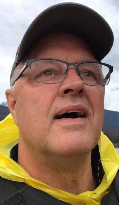 A close-up face shot of a man probably in his 50s. He's looking off to the right of the frame, wearing glasses and a baseball cap. He has a yellow piece of plastic tied around his neck while on a river outing. 