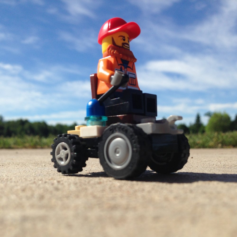A professional-quality photo shows a Lego "mayor" in a four-wheeled "wheelchair" driving on the pavement, with trees behind and a blue sky overhead. The photo is taken from an angle below that makes the Lego man appear life-size.